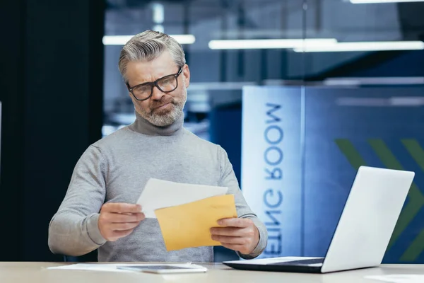 Mature gray-haired businessman inside modern office received letter message, senior man upset opens postal envelope, boss reads bad news upset and sad working at desk with laptop.