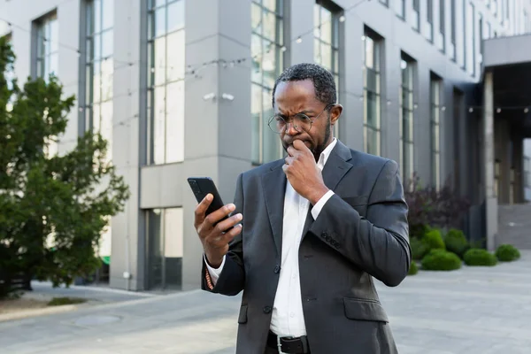 Mature senior african american boss reading bad news on phone, businessman in business suit outside office building using smartphone upset and disappointed by notification.