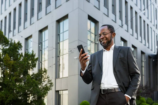 Successful and satisfied african american businessman using phone outside modern office building, mature man in business suit smiling and looking at smartphone screen texting.