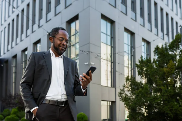 Başarılı ve tatmin olmuş Afro-Amerikan iş adamı modern ofis binasının dışında telefonu kullanıyor, takım elbiseli olgun bir adam gülümsüyor ve akıllı telefon ekranındaki mesajlara bakıyor..