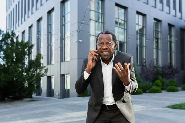 Angry african american boss arguing and shouting while talking on the phone, senior man in business suit outside office building.