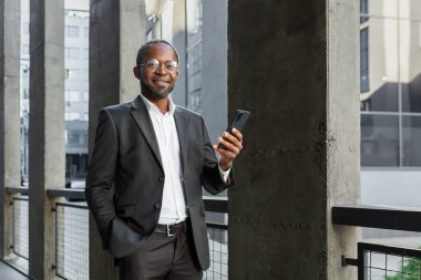 Afro-Amerikalı yetişkin iş adamı portresi, ofis binasının dışındaki yaşlı adam elinde telefonla gülümseyen ve kameraya bakan..
