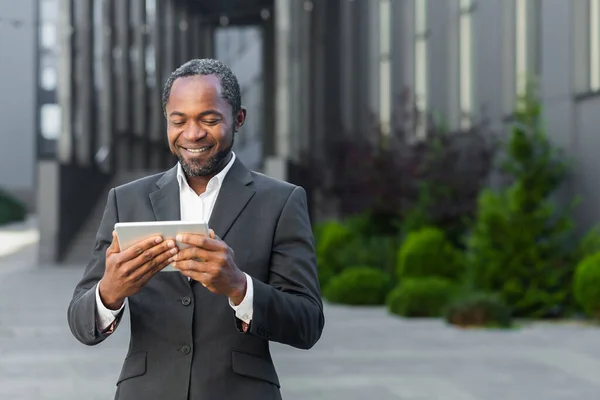 African american man in business suit outside office building watching online video with using app on tablet computer, businessman smiling and happy.