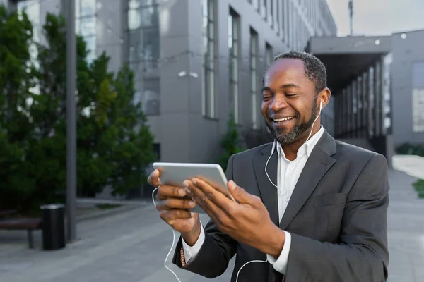African american man in business suit outside office building watching online video with using app on tablet computer, businessman smiling and happy.