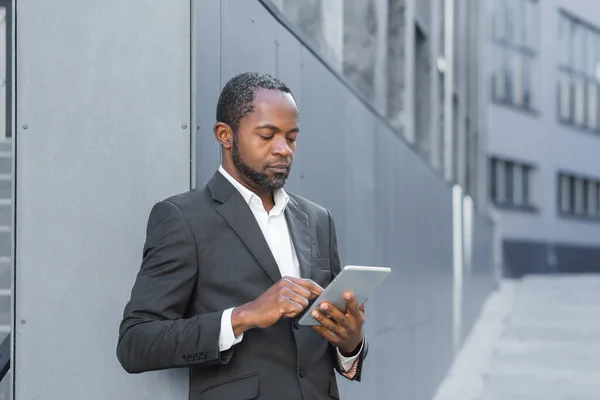 Serious thinking african american businessman using tablet computer, man in business suit mature reading news online standing outside office building.