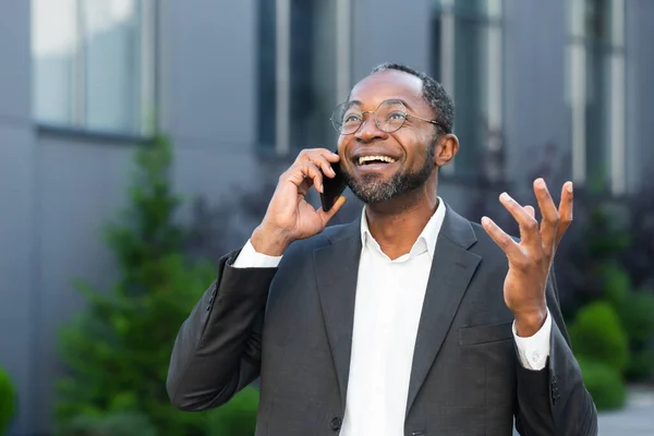 Cheerful and satisfied african american boss outside office building smiling and talking to colleagues on phone, man in business suit walking on lunch break.