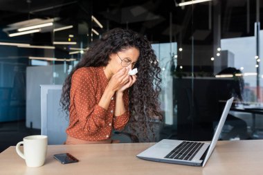 Ofiste çalışan hasta bir kadın, soğuk algınlığı geçiren, sıcak çay içen, hapşıran, iş yerinde öksüren, peçete kullanan, dizüstü bilgisayarla aynı masada oturan İspanyol bir kadın..