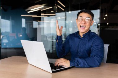 Başarılı bir Asyalı işadamı zafer başarısını kutluyor, ofiste sıradan gömlekli bir adamın portresi, çalışanın gülümsemesi ve zafer jestini kaldırarak elde ettiği başarıdan memnun olan kameraya bakması..