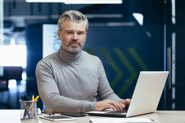 Portrait of serious gray haired boss, man looking at camera thinking, mature businessman inside office working at desk using laptop at work.
