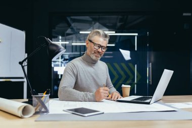 Ofiste başarılı bir kıdemli mimar, olgun gri saçlı bir adam plan çiziyor, gülümsüyor ve işten memnun..