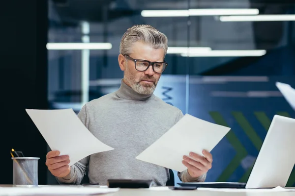 Frustrated and angry businessman with documents, mature gray businessman checking financial reports, boss not satisfied with achievement results and income working inside office with laptop.