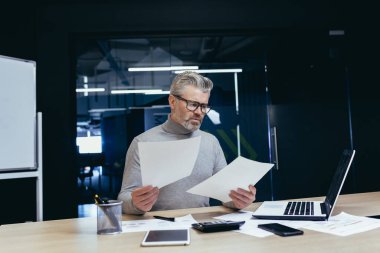 Belgelerle hayal kırıklığına uğramış ve kızgın iş adamı, finansal raporları kontrol eden olgun gri işadamı, patron başarı sonuçlarından memnun değil ve ofis içinde dizüstü bilgisayarla çalışan gelir..