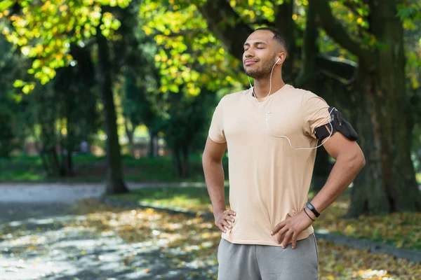 Latin american sportsman resting after jogging in the park and exercising, closed his eyes and listens to music and online audio books and podcast uses phone and headphones smiling