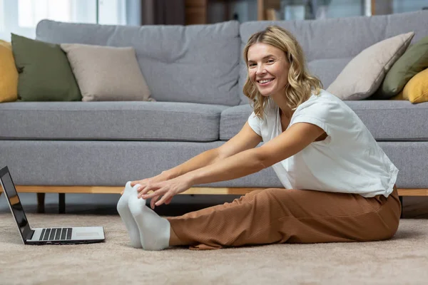 Portrait of active woman at home doing fitness and exercise and yoga, mature housewife looking at camera and smiling using laptop for online fitness training with trainer.