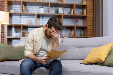 Upset man at home sitting on sofa reading letter with bad news notification in envelope opening, unhappy in living room and sad.