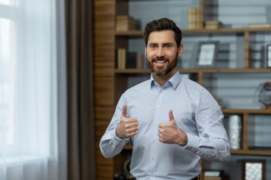 Successful businessman smiling and looking at camera, man shows thumbs up affirmatively and joyfully agrees with achievement, boss in shirt inside office at work.