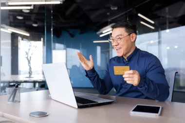 A happy young Asian man is sitting in the office at the table with a laptop, holding a credit card in his hand. Online payment, online shopping, money transfer, account verification.