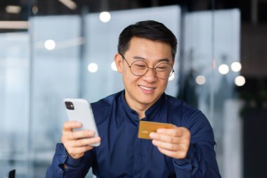 Close up photo. A happy young Asian man is sitting in the office at the table, holding a credit card and phone in his hands. Online payment, online shopping, money transfer, account verification.