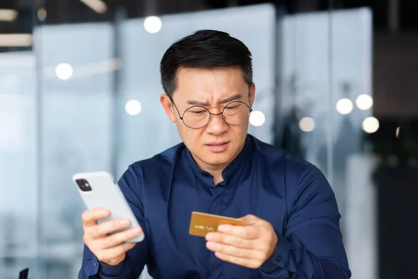 A worried young man is sitting in the office, holding a credit card and phone in his hands. Unsuccessful online shopping, transaction, lack of money in the account, blocked account. bankruptcy.