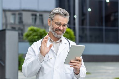 Senior gray-haired smiling doctor talking to patients remotely, a man in a medical coat and a stethoscope holds a tablet computer in his hands, consults patients online from outside a modern clinic.