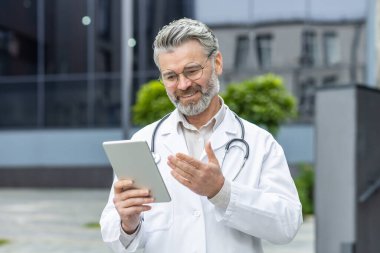 Senior gray-haired smiling doctor talking to patients remotely, a man in a medical coat and a stethoscope holds a tablet computer in his hands, consults patients online from outside a modern clinic.