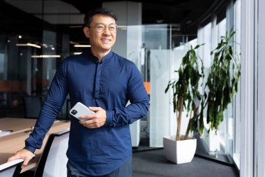 Portrait of a confident smiling Asian man. Businessman, director. He is standing in the office at the table, holding the chair with his hand, holding the phone in his hand, looking out the window.