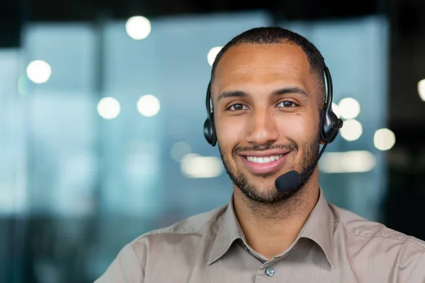Close up hispanic male worker with video headset smiling and looking at camera, tech support worker working inside office.