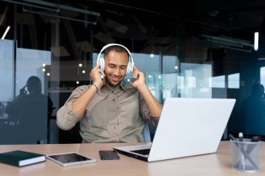 Successful young african american businessman relaxing in office, man in shirt and headphones listening to music online radio and podcasts, sitting at desk using laptop at work.