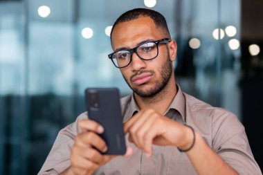 Serious thinking businessman close up, man holding smartphone and reading news online, worker inside office near window in shirt and glasses using phone typing message.