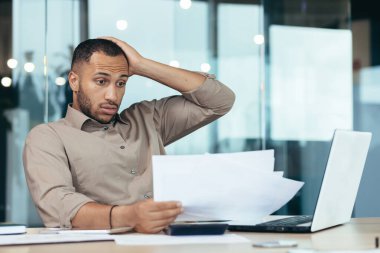 Pensive serious businessman reading financial report, hispanic businessman holding document in hands looking disappointed, working inside modern office with laptop behind paper work.