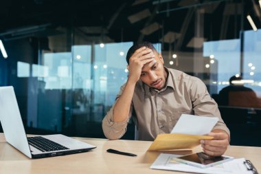 Pensive businessman inside office reading notification letter, man received envelope thinking about decision sitting at table and laptop inside office, bad news message.