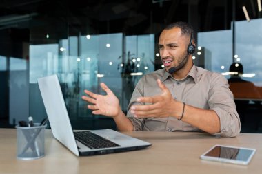 Angry worker with video call headset talking to customer, hispanic man explaining angry and upset, businessman inside modern office with laptop for customer service support tech.