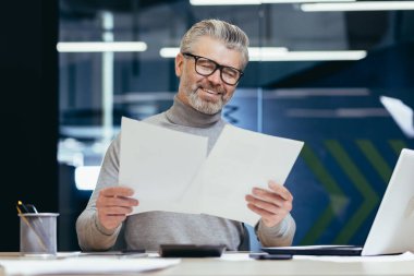 Senior smiling gray-haired businessman working in office at desk with laptop and documents. Holds in his hands, looks at sheets of paper, agreements, bills.