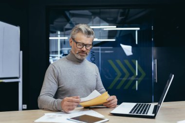 Senior gray-haired man working in office at desk with laptop and documents. He is holding an envelope with a letter, bills, and reports.