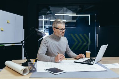 Senior gray-haired man architect, engineer, designer sits in the office at the table, works on a laptop, drawings. Makes notes.