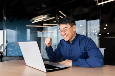 A happy young Asian man sits in the office at the table, looks at the laptop, talks on a video call, online meeting, rejoices at the success, the deal, the win.