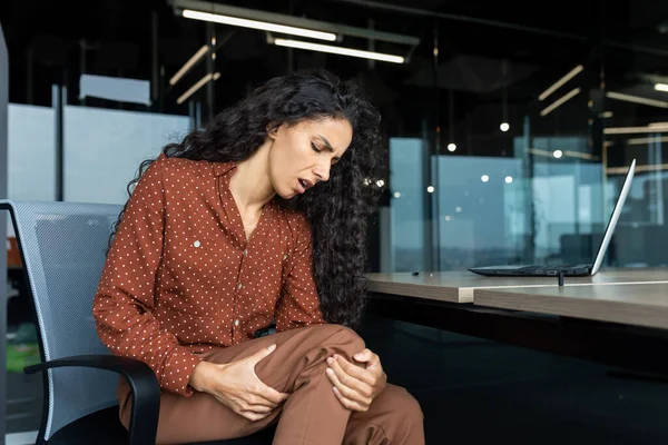 Latin American woman working inside office, business woman has severe leg pain, massaging muscle while sitting at table on chair, using laptop at work.