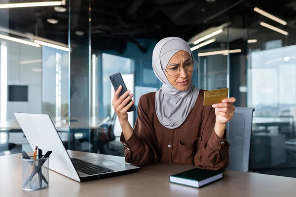 Upset and disappointed woman in hijab inside office trying to make purchase in online store, business woman holding smartphone and bank credit card, received money transfer error.