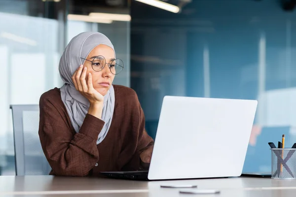 Serious bored businesswoman inside office, muslim woman in hijab thinking while sitting at workplace with laptop, woman at work thinking about decisions.