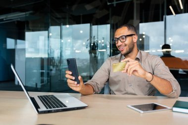 Successful hispanic businessman inside office, man in shirt smiling and happy holding bank credit card and phone, making money transfer and online shopping.