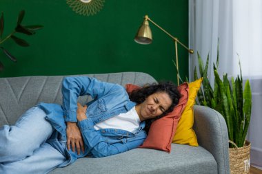 Hispanic woman sick lying on sofa in living room, woman has severe stomach pain, upset holding hands to side of stomach.