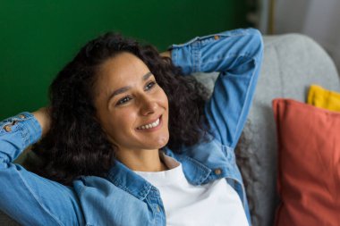 Beautiful hispanic woman daydreaming sitting on couch at home and looking out window, happy woman with hands behind head close up.