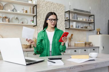 Angry young woman sitting in the kitchen at home using a laptop. Holds documents, a letter in his hands, types on the phone.