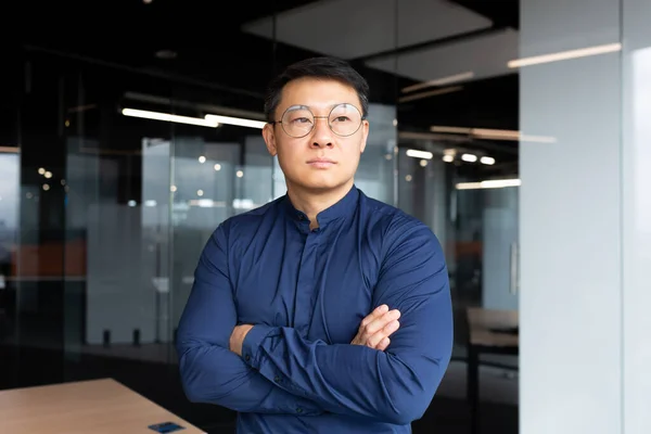 Asian serious and thinking man inside office looking out the window, businessman with crossed arms in shirt and sunglasses, boss thinking about an important decision.