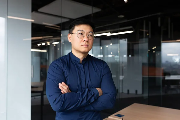 Asian serious and thinking man inside office looking out the window, businessman with crossed arms in shirt and sunglasses, boss thinking about an important decision.