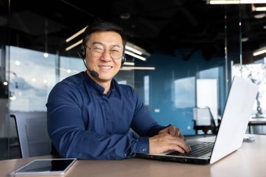 Portrait of successful office worker with headset for video call, asian man smiling and looking at camera working inside office, man in shirt and glasses customer service support.