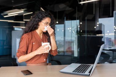 Sick young hispanic woman sitting at a desk in an office using a laptop. He wipes his nose with a napkin, has a runny nose, feels bad, holds a cup, drinks medicine, a hot drink.
