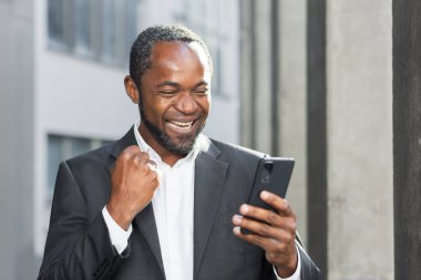 Successful african american boss outside office building using phone businessman in suit celebrating victory, successful achievement, man holding hand up triumph gesture, reading online notification