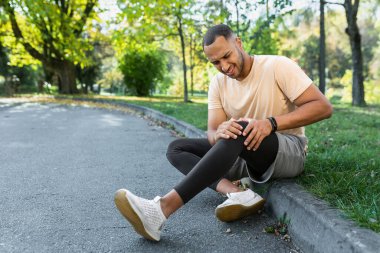 Man injured leg while jogging, african american man sitting on ground, massaging sore muscle with hands, upset and injured runner. clipart