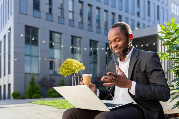 African american businessman outside office building talking to colleagues remotely, boss in business suit with laptop having video call, smiling with headphones headset sitting on bench.